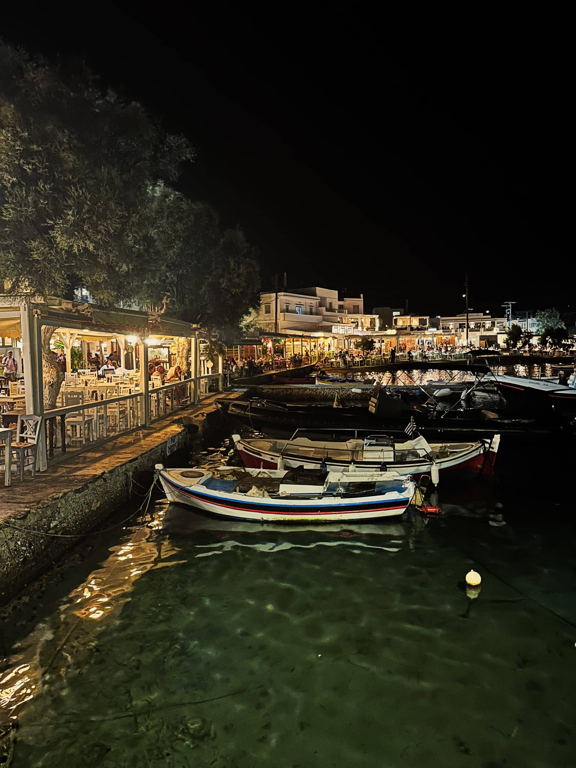 A nighttime picture of restaurants along the water with small boats in the bay. 