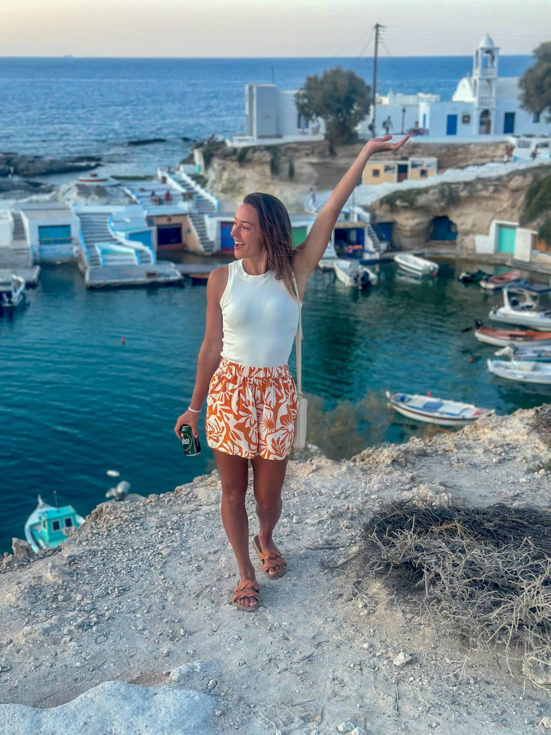 Kait smiling with arm out. She is standing on a cliff overlooking the small boat harbor in Madrakia, filled with colorful boathouses.