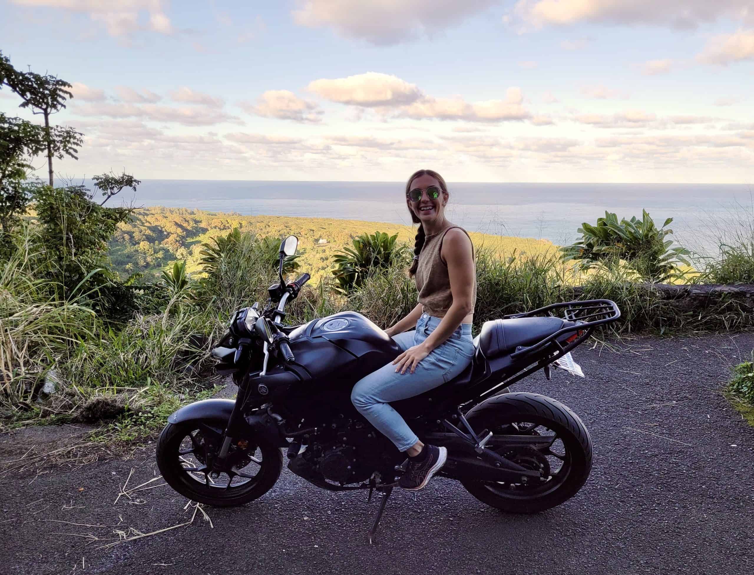 Kait sitting on a black motorcycle overlooking lush green hills and the ocean in the distance.
