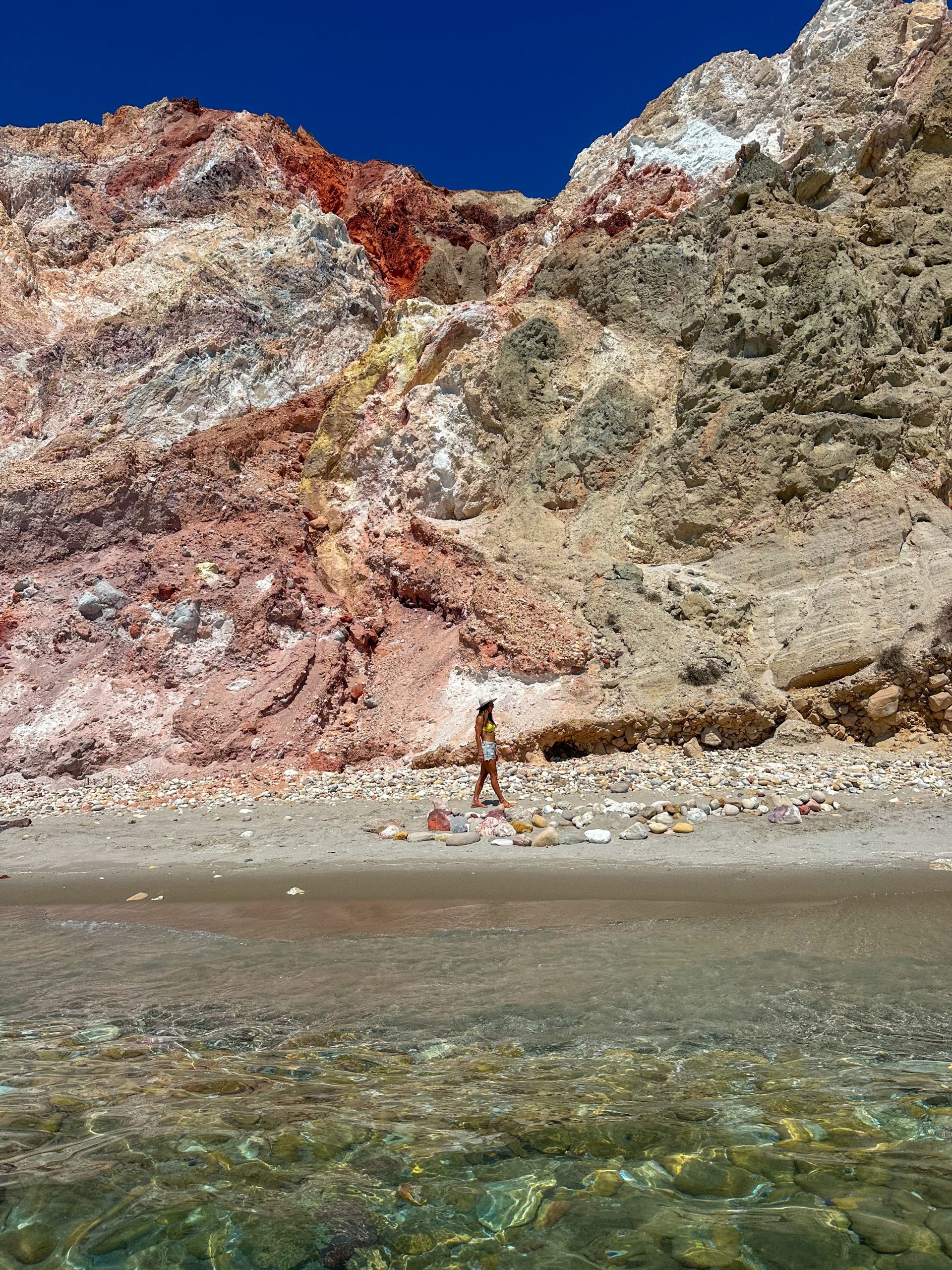 Kait walking along the beach in Milos beneath dramatic volcanic cliffs with vibrant red, yellow, and white colors.