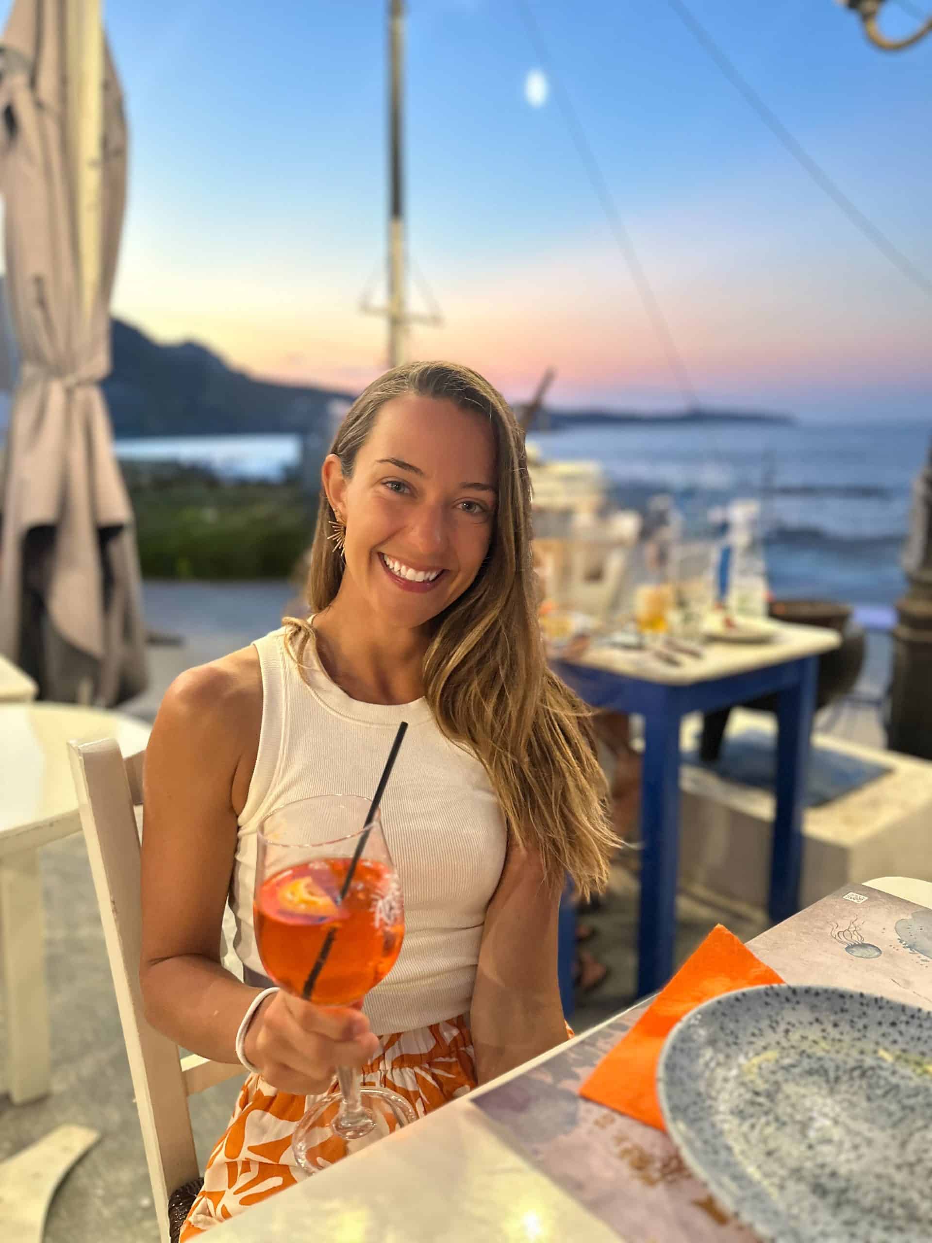 Kait enjoying an Aperol Spritz during a seaside sunset dinner in Milos Greece, overlooking the Aegean Sea.