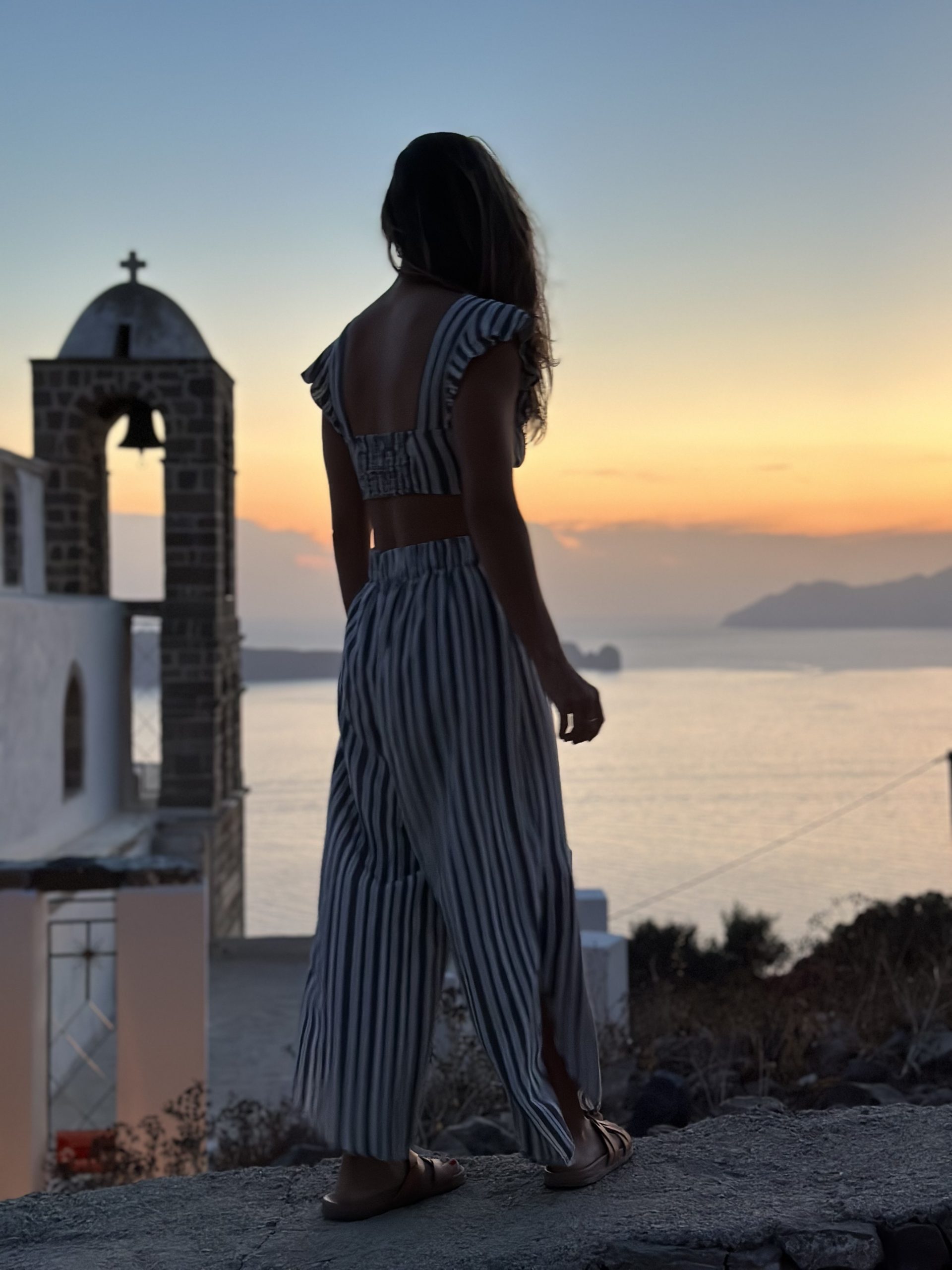 Kait looking out at the sunset from Plaka Castle in Milos, with a bell tower and sea views in the background.