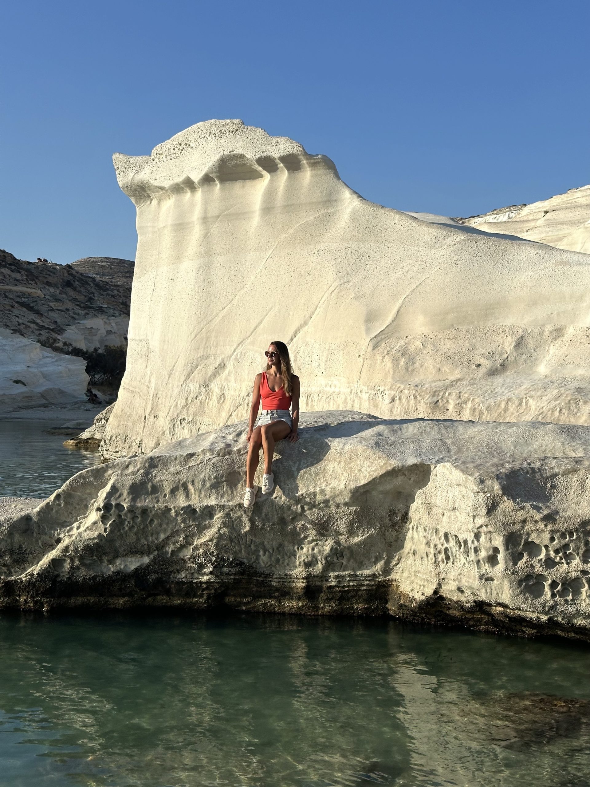 Kait sitting on the white volcanic cliffs of Sarakiniko Beach in Milos, surrounded by turquoise water and clear skies.
