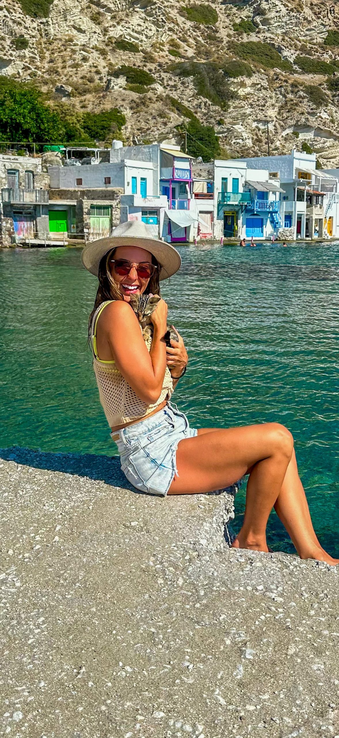 Kait sitting by the waterfront in Klima, Milos, holding a kitten with colorful syrmata houses in the background.
